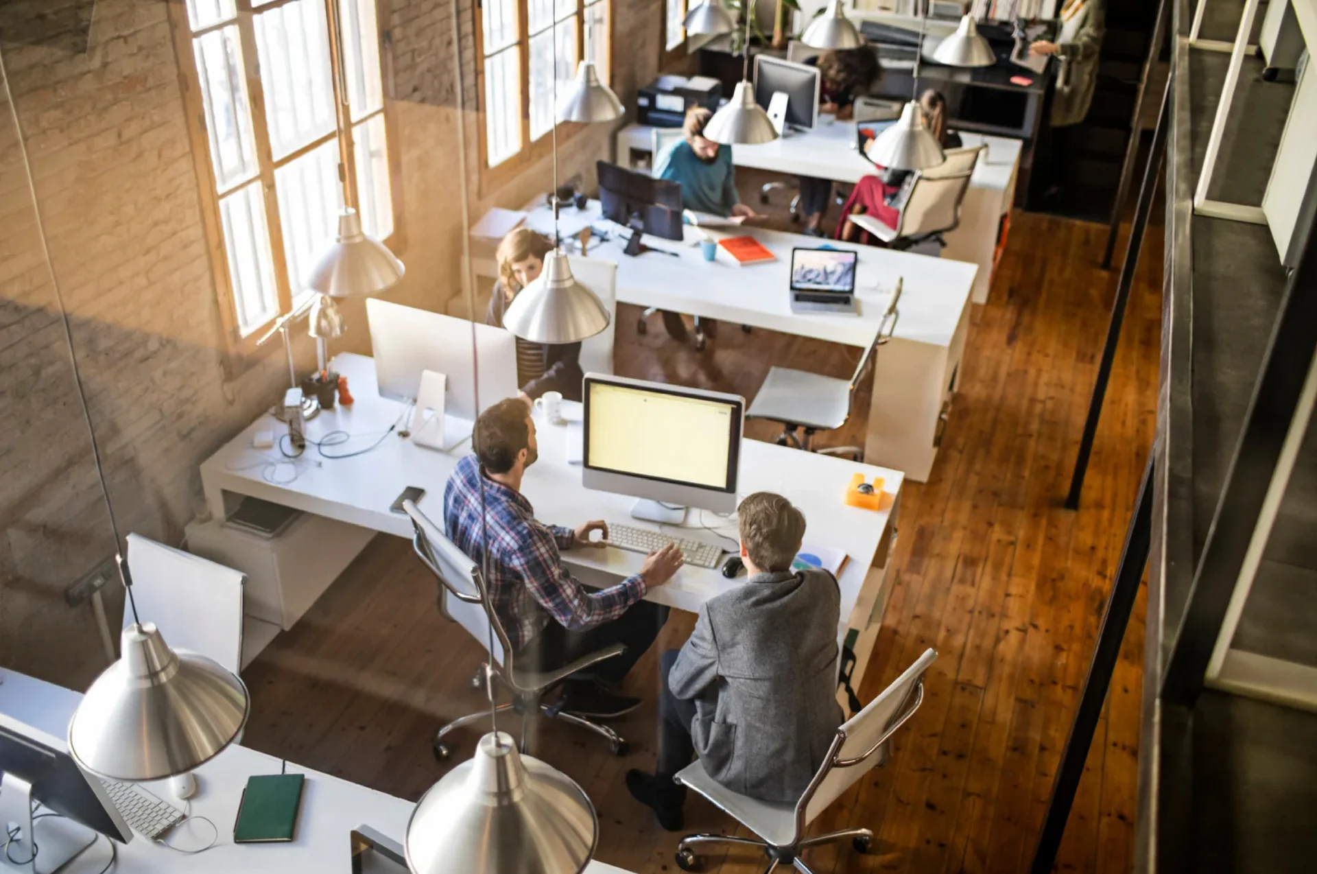 A modern open office viewed from above with multiple desks, computers, and people working at their workstations under natural light.