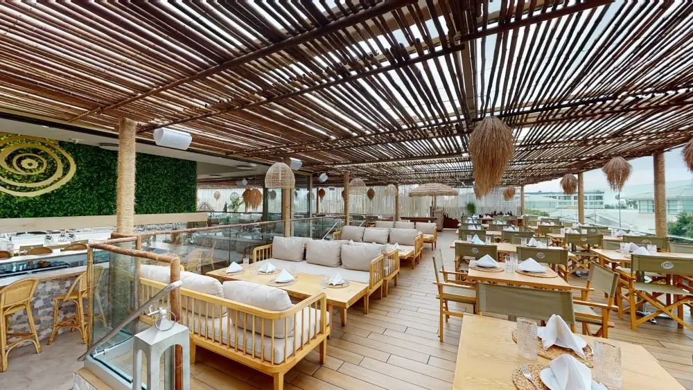 Outdoor cafe lounge with wooden pergola roof and hanging decorations, featuring cushioned wooden benches, tables, and chairs, with a moss wall art and views across the city